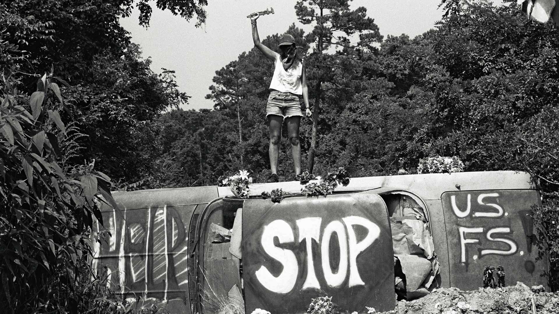 Greyscale picture of an activist standing on a tilted car with the words "Help" "Stop" and "US FS" painted on the car