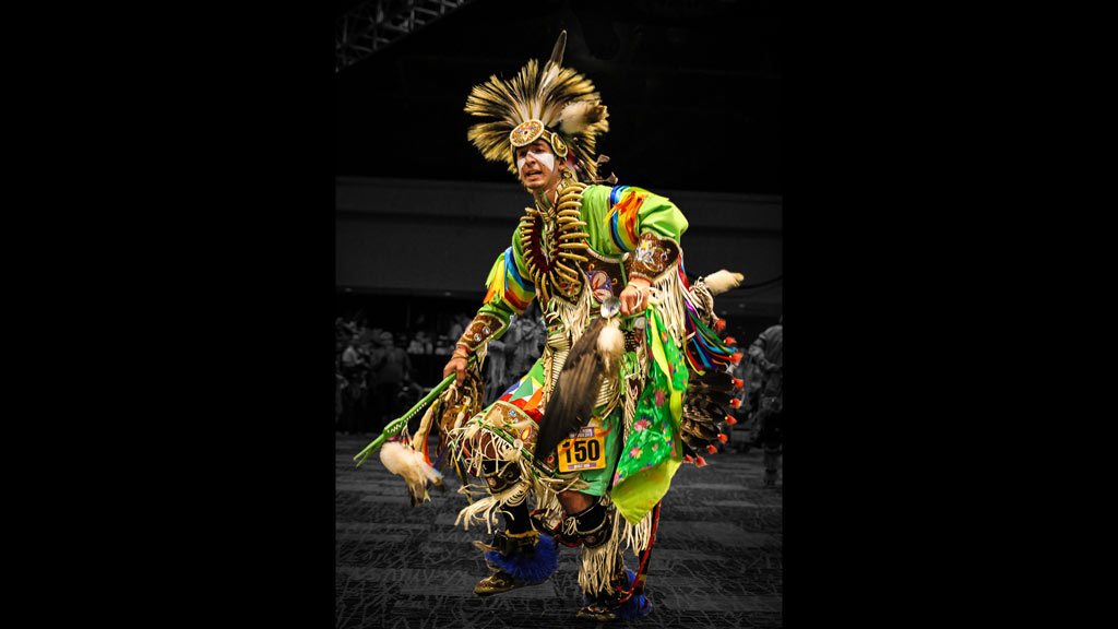 A man wearing bright green traditional powwow clothing while dancing in a conference room.