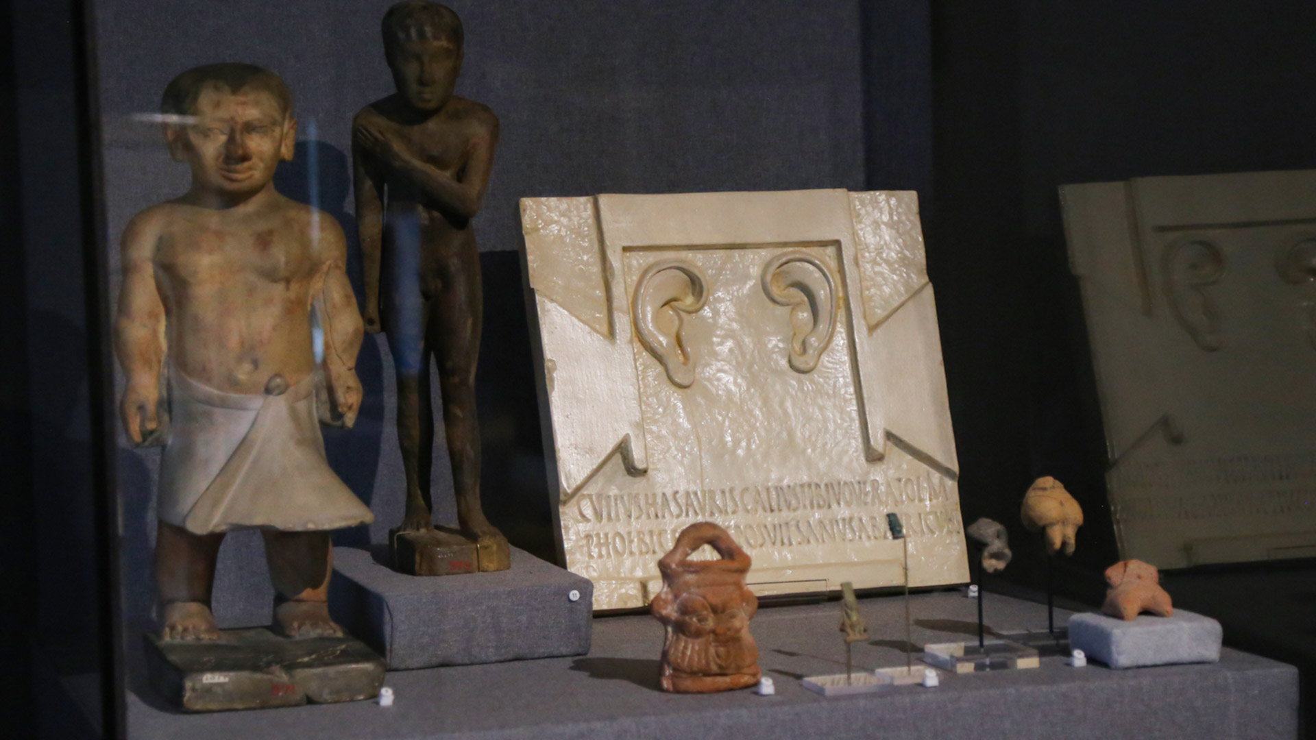 case shot including statues, figurines of the Goddess Bes, and a tablet depicting two ears with latin inscripted below