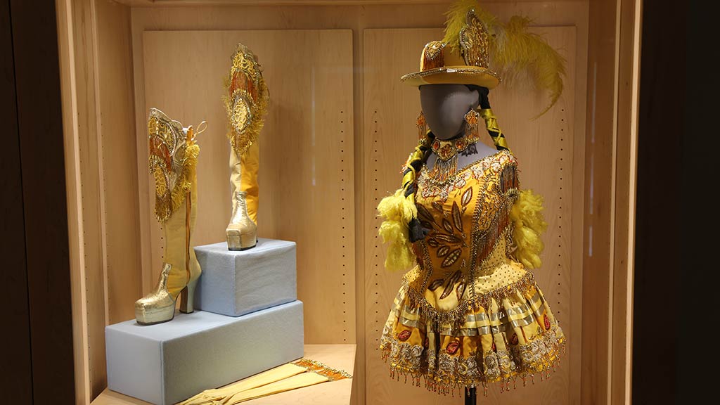 a model in costume that is various shades of yellow, rims of round gold beads, lace-like ribbon sewn onto the yellow fabric