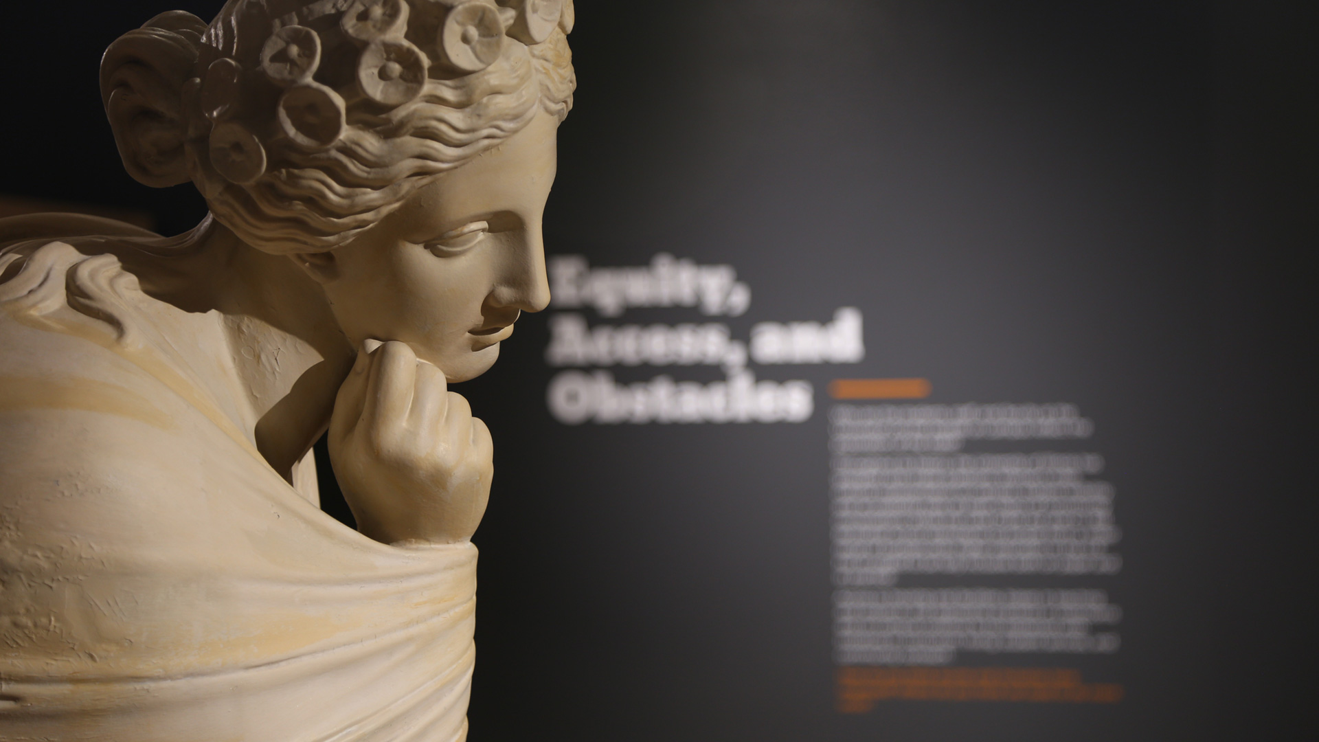 close-up of a statue with heading text in the background on a wall, reading 'equality, access, and obstacles'